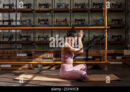 Woman in yoga pose on a mat with sewing machines. Sunlight and vintage machines create a calm, nostalgic atmosphere enhancing the yoga practice’s sere Stock Photo