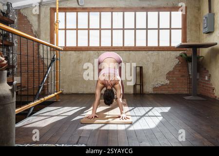 A woman practices yoga in a sunlit loft with rustic brick walls. She’s doing the downward dog pose in a serene, airy space, wearing comfortable pink s Stock Photo