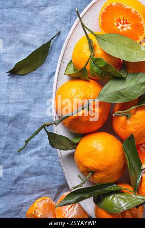 Fresh tangerines with vibrant green leaves are artfully arranged on a textured blue fabric, creating a striking contrast. A few are peeled, revealing Stock Photo