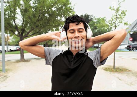 "Latino sportsman smiling as he listens to music with large headphones with eyes closed, stretching arms back on a sunny day in a park." Stock Photo