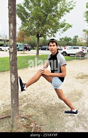 A Latino sportsman stretches his leg on a tree trunk in a park setting, dressed in athletic wear and sneakers, preparing for a run. Stock Photo
