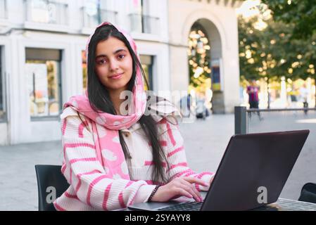 A arab student woman wearing a pink scarf and striped sweater is seated at an outdoor cafe, working on her laptop. The background contains a blurred c Stock Photo