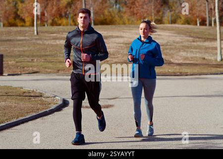 A young man and woman jog side by side in a park during autumn. They are dressed in athletic wear, enjoying a fitness routine amidst colorful fall fol Stock Photo