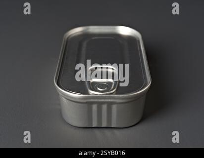 A detailed view of a metal food can featuring a pull tab on its lid. The can is placed on a smooth, dark gray surface, highlighting its shiny metallic Stock Photo