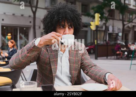 A mixed race man with curly hair teleworks from a street cafe, sipping coffee, dressed in a stylish jacket, capturing the lifestyle of remote workers. Stock Photo