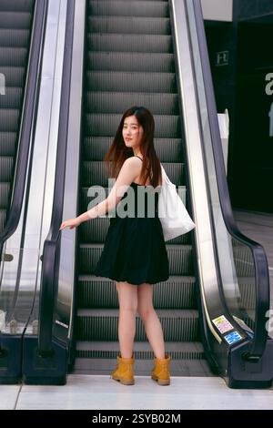 A young asian woman in a black dress and boots stands on an escalator in an urban setting, holding a tote bag. The scene captures a modern city lifest Stock Photo