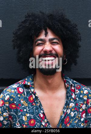 A man with curly hair laughs joyfully, wearing a vibrant floral shirt against a gray background. His expression radiates happiness and positivity, cap Stock Photo