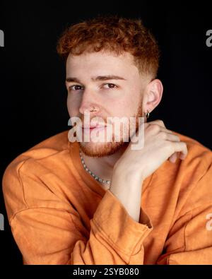 A young man with red hair and beard wearing an orange sweater poses against a dark background. He has a nose ring and small earring, exuding a confide Stock Photo