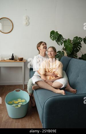 A mother and daughter share a joyful moment while engaging in a self-care routine at home. They are smiling and relaxed, sitting on a cozy sofa, surro Stock Photo