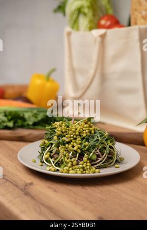 A fresh topped with green sprouts on a white plate sits on a wooden table. In the background, assorted vegetables and a reusable bag add a vibrant tou Stock Photo