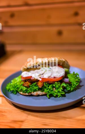Close-up of a Greek-style sandwich with fresh lettuce, tomatoes, seasoned meat, and creamy sauce on a gray plate. Served on a wooden table, perfect fo Stock Photo