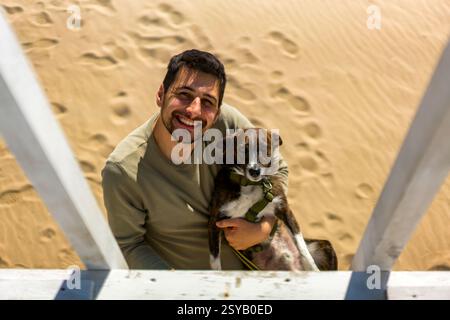 A cheerful man holding his dog on a sunny beach in Greece. The sand is marked with footprints. Captures joy and companionship in a serene coastal sett Stock Photo
