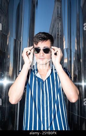 Fashionable young man in a striped shirt adjusting his sunglasses while posing against a reflective metal background in the city. Confident, urban, an Stock Photo