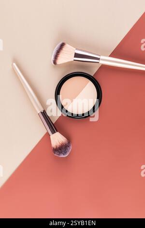 A pair of makeup brushes and a compact powder are artistically arranged on a split tone background in beige and rust, highlighting a minimalist aesthe Stock Photo