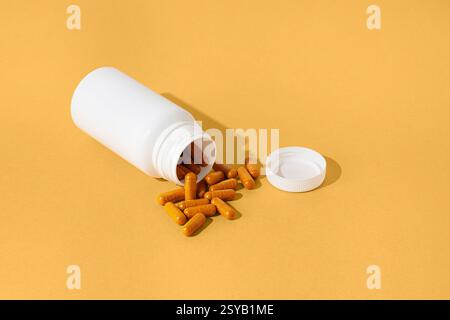A white plastic bottle lies open on a vibrant yellow surface, spilling out orange capsules. The minimalist composition highlights the capsules vivid c Stock Photo