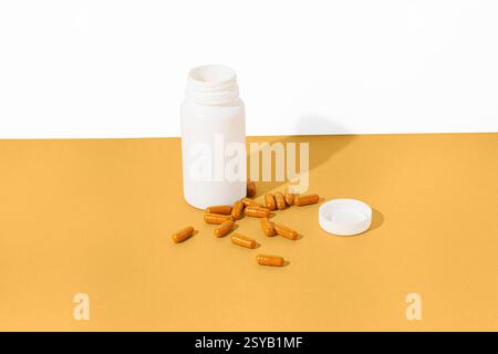 A white bottle with its lid off lies on a yellow surface, surrounded by scattered orange capsules. The bright, minimalist setup emphasizes the contras Stock Photo