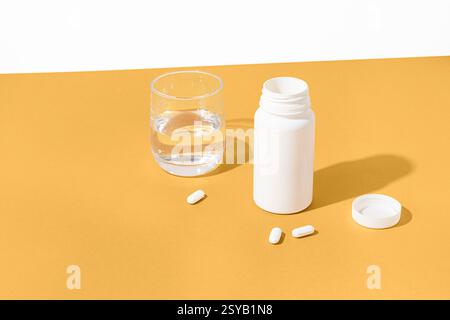 A white pill bottle with scattered pills beside a glass of water on a vibrant yellow background. The open bottle cap lies nearby, illustrating a healt Stock Photo