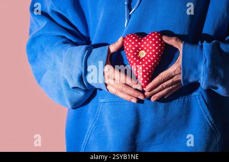 Close up of a person in a blue hoodie holding a red heart shaped pillow with white polka dots and a wooden button, symbolizing love yourself and comfo Stock Photo