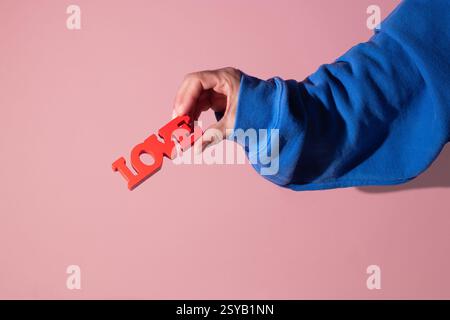 A hand in a blue sleeve holds a red love sign against a soft pink background, creating a vibrant and emotionally resonant image perfect for themes of Stock Photo
