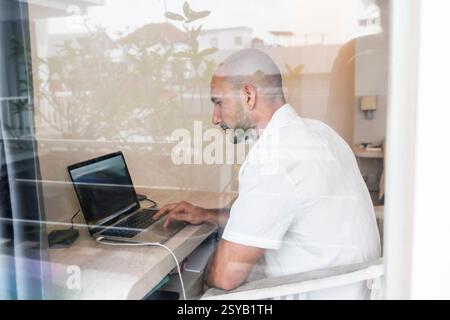 A man sits at a desk in a hotel room, focused on his laptop screen. Through reflections in the window, an image of concentration and modern work cultu Stock Photo