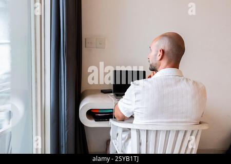 A man seated at a desk in a hotel room, working on a laptop. The bright interior and modern furnishings create a productive environment for remote wor Stock Photo