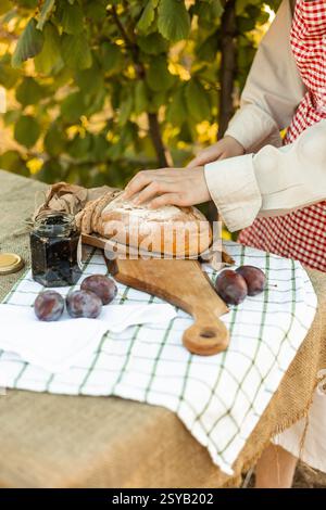 A person slices fresh bread on a wooden board outdoors, surrounded by jars and plums. The rustic setting highlights the charm of making canapes in nat Stock Photo