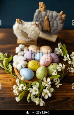 A rustic setting featuring colorful speckled Easter eggs in a wooden bowl, surrounded by white blossoms and woolly chickens decor on a wooden table. Stock Photo