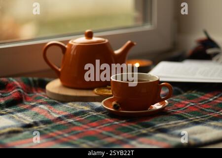 A warm setting featuring a orange teapot and cup on a plaid blanket near a window. A lit candle and an open book add to the cozy, inviting ambiance. Stock Photo