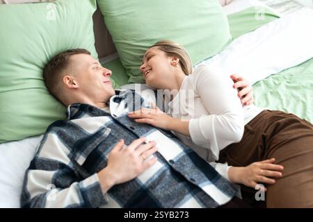 A young man and woman lie comfortably on a bed, sharing a close, intimate moment. They smile at each other, expressing affection and happiness in a co Stock Photo