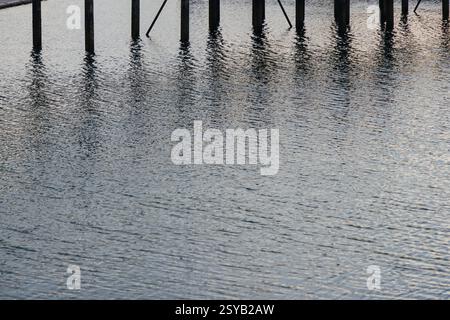 Water reflections with wooden poles standing vertically. The ripples and light reflections create a calm and textured scene. Stock Photo