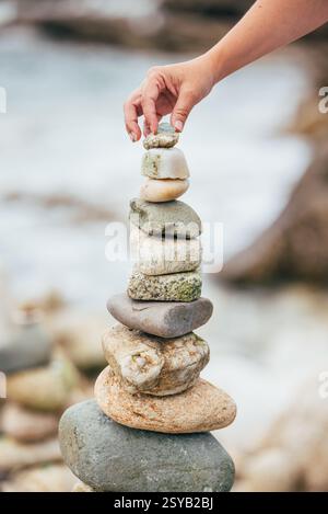 A hand carefully places a small stone atop a balanced stack of larger rocks by the seaside. The focus is on the stones and the hand, with the sea soft Stock Photo
