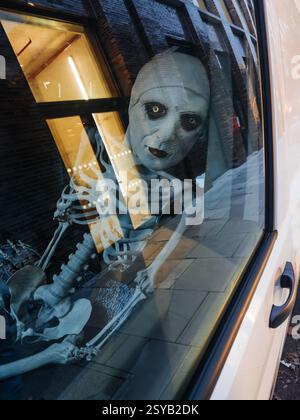 A haunting skeleton figure peers through a city window, its eerie reflection captured on the glass. The scene blends urban architecture with Halloween Stock Photo