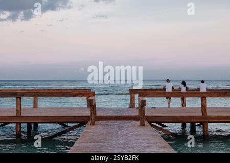 A tranquil scene at Tulum, Riviera Maya, featuring anonymous people relaxing on a wooden pier as the sun sets over the ocean. The calm waters and past Stock Photo