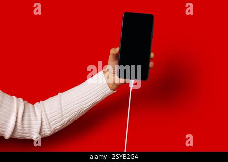 Side view of unrecognizable person in casual clothes holding smartphone with black screen while standing against red background Stock Photo