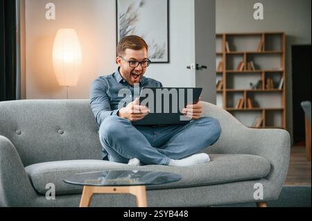 Celebrating success. Male freelance worker is using laptop at home. Cozy apartment. Stock Photo