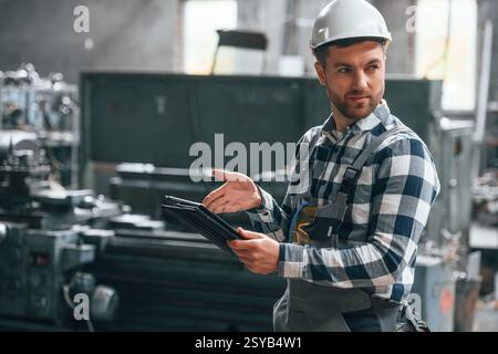 Standing and using graphic tablet. Factory male worker in uniform is indoors. Stock Photo