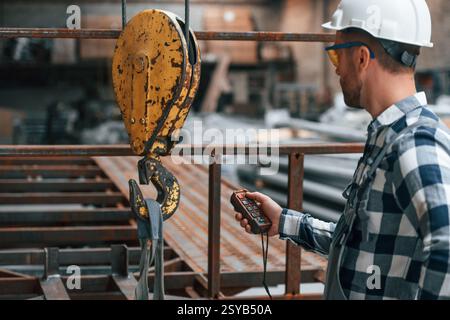 Using the controller with buttons to operate the little crane machine. Factory male worker in uniform is indoors. Stock Photo