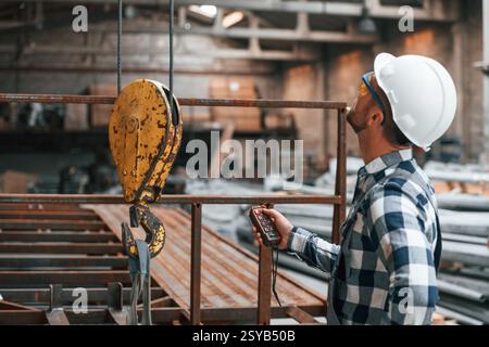 Using the controller with buttons to operate the little crane machine. Factory male worker in uniform is indoors. Stock Photo