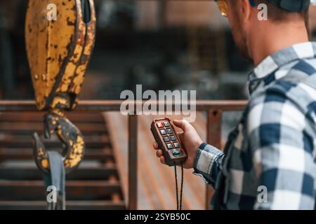 Using the controller with buttons to operate the little crane machine. Factory male worker in uniform is indoors. Stock Photo
