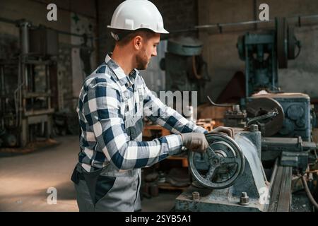 Side view. Using machine. Factory male worker in uniform is indoors. Stock Photo