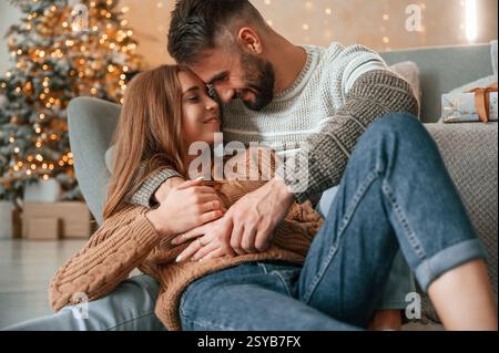 Laying down on the sofa and embracing. Lovely young couple are celebrating New Year at home. Stock Photo
