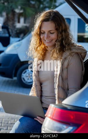 Freelancer woman sitting in the open trunk of a car using laptop Stock Photo