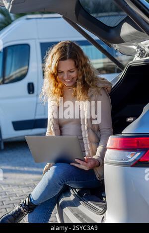 Freelancer woman sitting in the open trunk of a car using laptop Stock Photo