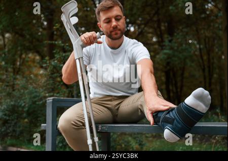 Sitting, touching the leg. Man with crutches is in the park outdoors. Stock Photo