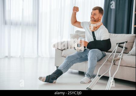 Sitting, holding soccer ball, watching sport match. Man with crutches is at home indoors. Having injuries. Stock Photo