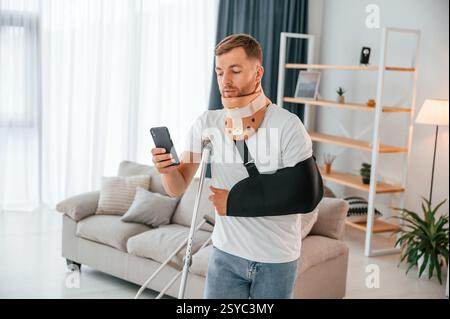 Using smartphone. Man with crutches is at home indoors. Having injuries. Stock Photo