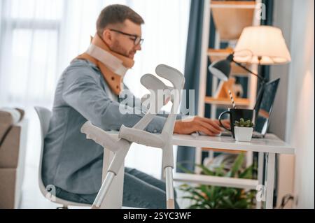Working from home by using laptop. Man with crutches is indoors. Having injuries. Stock Photo