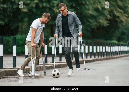 Having fun, playing soccer. Two men with crutches is outdoors on the road. Stock Photo
