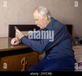 Elderly Man Sitting on Bed, Leaning on Table, Looking at Phone Stock Photo