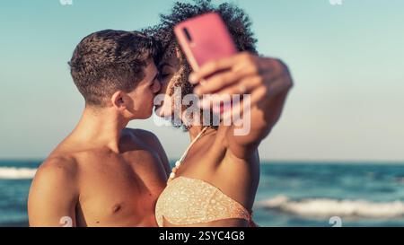 Young interracial couple kissing while taking a selfie on the beach during summer. Romantic and spontaneous moment capturing love and happiness by the Stock Photo